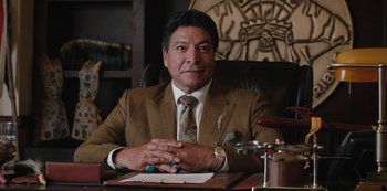 Movie still from “Yellowstone” (2018), created by John Linson – A man sitting at a desk wearing a suit and tie; Medium shot, Low angle
