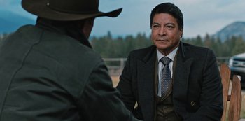 Movie still from “Yellowstone” (2018), created by John Linson – A man wearing a suit and tie talking to another man wearing a hat; Medium shot, Over the shoulder angle