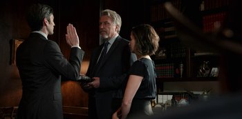 Movie still from “Yellowstone” (2018), created by John Linson – A man in a suit is being sworn in by a woman in a black dress; Medium shot, Over the shoulder angle