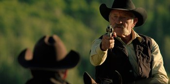 Movie still from “Yellowstone” (2018), created by John Linson – An old man in a cowboy hat pointing a gun at another man; Medium shot, Over the shoulder angle
