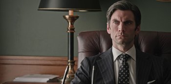 Movie still from “Yellowstone” (2018), created by John Linson – A man in a suit sitting in a chair in front of a lamp; Close Up shot, Low angle