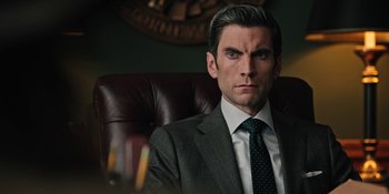 Movie still from “Yellowstone” (2018), created by John Linson – A man in a suit and tie sitting in a chair; Close Up shot, Over the shoulder angle