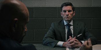 Movie still from “Yellowstone” (2018), created by John Linson – A man in a suit sitting at a table with his hands crossed; Close Up shot, Over the shoulder angle