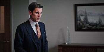 Movie still from “Yellowstone” (2018), created by John Linson – A man in a suit and tie standing next to a table; Close Up shot, Low angle