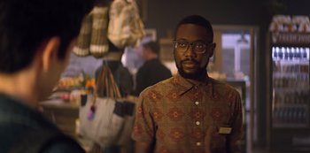 Movie still from “You” (2018), created by Sera Gamble – A man wearing glasses standing in front of a group of people; Medium shot, Over the shoulder angle