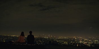 Movie still from “You” (2018), created by Sera Gamble – A man sitting on top of a hill at night with a city in the background; Extreme Wide shot, Low angle