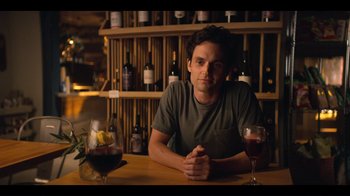 Movie still from “You” (2018), created by Sera Gamble – A man sitting at a table in front of a glass of wine; Medium shot, Over the shoulder angle