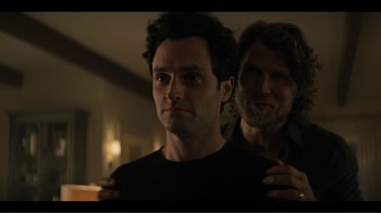 Movie still from “You” (2018), created by Sera Gamble – A man standing next to another man in a dark room; Medium shot, Over the shoulder angle
