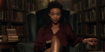 Movie still from “You” (2018), created by Sera Gamble – A woman sitting in a chair holding a book in her lap; Medium shot, Low angle