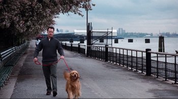 Movie still from “You've Got Mail” (1998), directed by Nora Ephron – A man walking a dog on a leash on a sidewalk near a body of water; Wide shot, Low angle