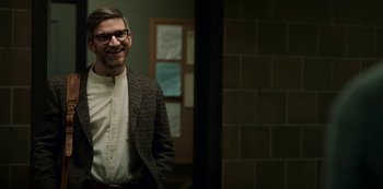 Movie still from “Archive 81” (2022), created by Rebecca Sonnenshine – A man wearing a suit and glasses smiling for the camera; Medium shot, Over the shoulder angle