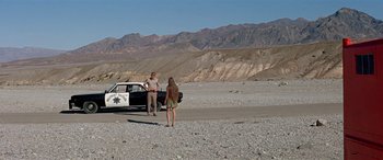 Movie still from “Zabriskie Point” (1970), directed by Michelangelo Antonioni – A man and a woman standing in front of a police car; Wide shot, Low angle