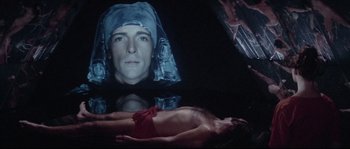 Movie still from “Zardoz” (1974), directed by John Boorman – A man laying on the ground in front of an image of a woman; Medium shot, Overhead angle