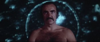 Movie still from “Zardoz” (1974), directed by John Boorman – A man with no shirt is standing in front of an image of the moon; Medium shot, Low angle