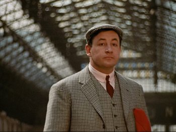 Movie still from “Zazie dans le Métro” (1960), directed by Louis Malle – A man in a suit and tie standing in a train station; Medium shot, Low angle