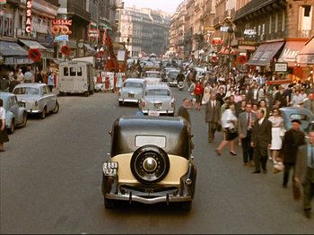 Movie still from “Zazie dans le Métro” (1960), directed by Louis Malle – An old car driving down a busy city street; Extreme Wide shot, High angle