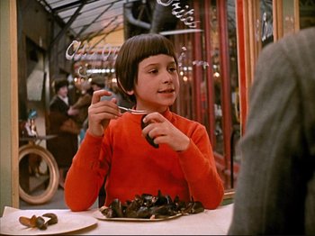 Movie still from “Zazie dans le Métro” (1960), directed by Louis Malle – A young girl sitting at a table with a plate of food in front of her; Medium shot, Over the shoulder angle