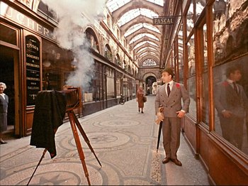 Movie still from “Zazie dans le Métro” (1960), directed by Louis Malle – A man in a gray suit and a white hat is standing in front of an open window with smoke coming out of it; Wide shot, High angle