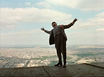 Movie still from “Zazie dans le Métro” (1960), directed by Louis Malle – A man standing on the edge of a building with his arms outstretched; Extreme Wide shot, Low angle