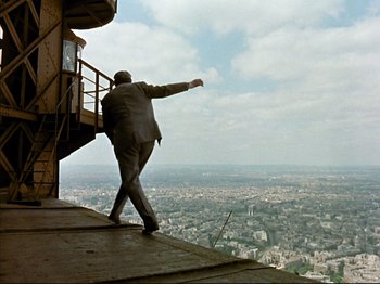 Movie still from “Zazie dans le Métro” (1960), directed by Louis Malle – A man standing on the side of a building near a city; Wide shot, Low angle