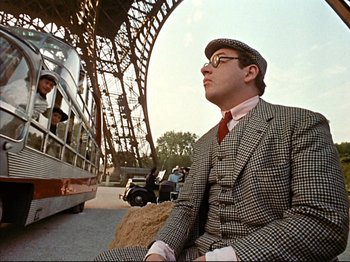 Movie still from “Zazie dans le Métro” (1960), directed by Louis Malle – A man sitting on a bench in front of an eiffel tower; Medium shot, Low angle