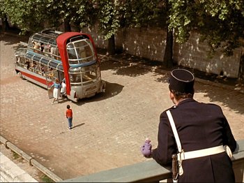 Movie still from “Zazie dans le Métro” (1960), directed by Louis Malle – A man standing on the side of a road next to a red and blue bus; Extreme Wide shot, High angle