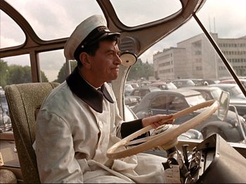 Movie still from “Zazie dans le Métro” (1960), directed by Louis Malle – A man in a uniform driving a car in a parking lot; Medium shot, High angle