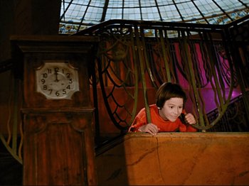 Movie still from “Zazie dans le Métro” (1960), directed by Louis Malle – A little boy sitting in a chair in a room; Wide shot, Low angle