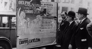 Movie still from “Zelig” (1983), directed by Woody Allen – A group of men standing next to a large poster; Medium shot, Low angle