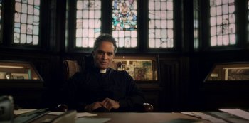 Movie still from “Archive 81” (2022), created by Rebecca Sonnenshine – A man sitting at a table in front of a stained glass window; Medium shot, Low angle