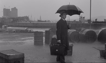 Movie still from “Zorba the Greek” (1964), directed by Michael Cacoyannis – A man holding an umbrella while standing in the rain; Wide shot, High angle