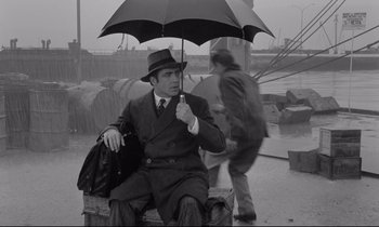 Movie still from “Zorba the Greek” (1964), directed by Michael Cacoyannis – A black and white photo of a man holding an umbrella; Wide shot, High angle
