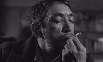 Movie still from “Zorba the Greek” (1964), directed by Michael Cacoyannis – A black and white photo of a man smoking a cigarette; Close Up shot, Low angle