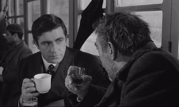 Movie still from “Zorba the Greek” (1964), directed by Michael Cacoyannis – Two men are sitting at a table and one of them is holding a drink; Medium shot, Over the shoulder angle