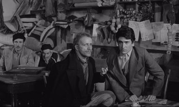 Movie still from “Zorba the Greek” (1964), directed by Michael Cacoyannis – A group of men sitting next to each other in a room; Medium shot, Over the shoulder angle