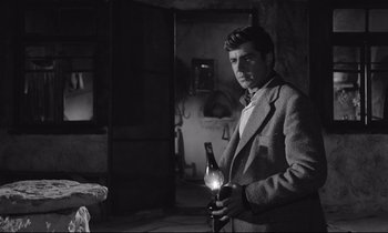 Movie still from “Zorba the Greek” (1964), directed by Michael Cacoyannis – A black and white photo of a man holding a bottle of wine; Medium shot, Low angle