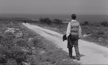 Movie still from “Zorba the Greek” (1964), directed by Michael Cacoyannis – A man walking down a dirt road near a body of water; Wide shot, Over the shoulder angle