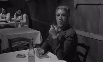 Movie still from “Zorba the Greek” (1964), directed by Michael Cacoyannis – Black and white photograph of a man sitting at a table; Medium shot, Low angle