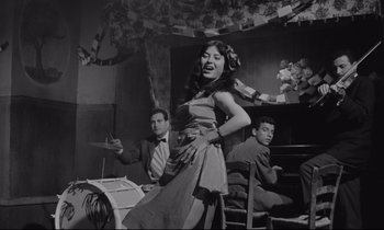 Movie still from “Zorba the Greek” (1964), directed by Michael Cacoyannis – A woman standing in front of two men in front of a piano; Medium shot, Low angle