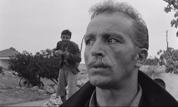 Movie still from “Zorba the Greek” (1964), directed by Michael Cacoyannis – A man standing next to another man on a dirt field; Close Up shot, Low angle