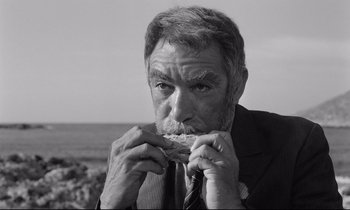 Movie still from “Zorba the Greek” (1964), directed by Michael Cacoyannis – An older man in a suit and tie eating a sandwich; Close Up shot, High angle