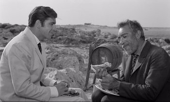 Movie still from “Zorba the Greek” (1964), directed by Michael Cacoyannis – Two men are sitting on a rocky hillside eating; Medium shot, Over the shoulder angle