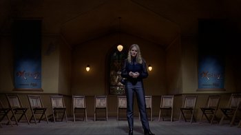 Movie still from “eXistenZ” (1999), directed by David Cronenberg – A woman standing in front of a row of chairs; Wide shot, Low angle
