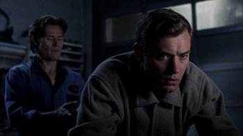Movie still from “eXistenZ” (1999), directed by David Cronenberg – A man sitting in front of another man in a dark room; Close Up shot, Over the shoulder angle
