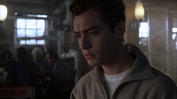 Movie still from “eXistenZ” (1999), directed by David Cronenberg – A young man looking at the camera while sitting at a table; Close Up shot, Over the shoulder angle