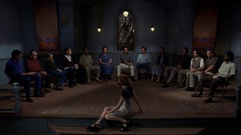 Movie still from “eXistenZ” (1999), directed by David Cronenberg – A group of people sitting in a circle on a stage; Wide shot, High angle