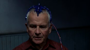 Movie still from “eXistenZ” (1999), directed by David Cronenberg – An older man wearing a blue star headband with a red string around his head; Close Up shot, Over the shoulder angle
