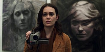 Movie still from “Archive 81” (2022), created by Rebecca Sonnenshine – A woman holding a video camera in front of a painting; Close Up shot, Over the shoulder angle