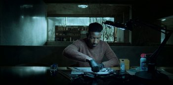 Movie still from “Archive 81” (2022), created by Rebecca Sonnenshine – A man sitting at a table cutting paper with scissors; Medium shot, Low angle