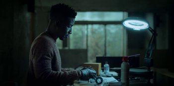 Movie still from “Archive 81” (2022), created by Rebecca Sonnenshine – A man sitting at a table looking at something in a jar; Medium shot, Low angle